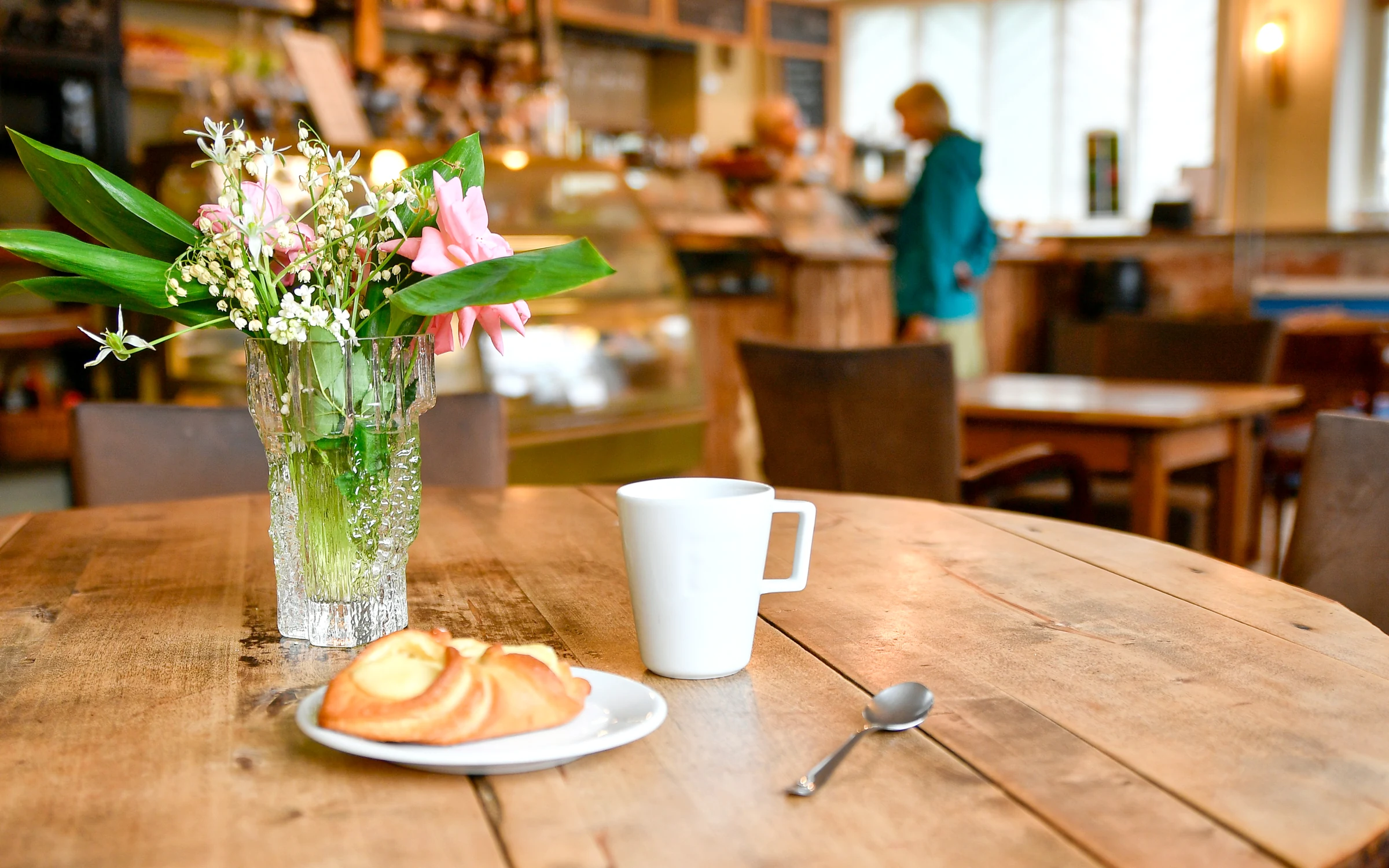 A pastry and a white mug of coffee beside a jug of fresh flowers on a worn oak table, with the café counter and big bright windows softly blurred behind.